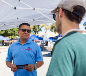 Jesse Armendarez holding a card speaking with a man under an easy-up