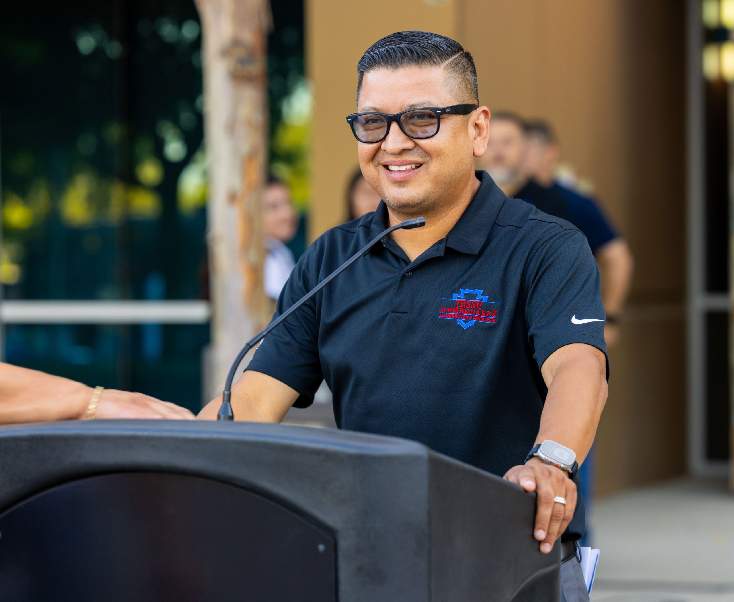 Jesse Armendarez standing and speaking at a podium with a building and people behind him.